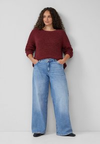 Weinroter Strickpullover mit weiten Ärmeln, kombiniert mit hellblauen weiten Jeans. Weiche Textur und lässige Passform, mit vorderen Taschen und Knopfverschluss.