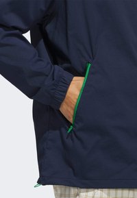 Chaqueta azul marino con puños elásticos y detalles de cremallera verde, que presenta un bolsillo lateral con una mano abierta. Tejido ligero con una textura suave.