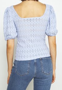 Světle modrý top s nafukovacími rukávy vyrobený z texturované látky s výřezy. Má čtvercový výstřih a přiléhavý střih. Kombinováno s džíny.