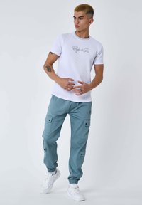 T-shirt bianco a coste con logo grigio. Pantaloni cargo azzurri con tasche a pattina e polsini elasticizzati. Sneakers bianche.