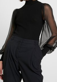 Maglione a collo alto nero a coste con maniche trasparenti e a sbuffo, abbinato a pantaloni a quadri in tonalità scure a vita alta. Texture morbide e forma strutturata.