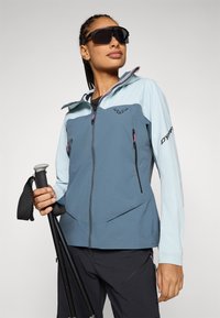 Lichte, twee-tone jas in blauw en lichtblauw, met ritsluiting, roze accenten en logo; vastgehouden met trekkingstokken in een zwarte grip.