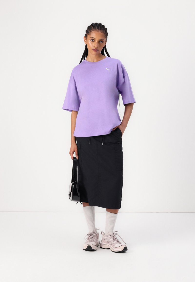 Lila übergroßes T-Shirt, schwarzer Mid-Length-Rock, weiße Sneaker mit rosafarbenen Akzenten und eine kleine schwarze Tasche, vor einem schlichten weißen Hintergrund.