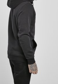 Schwarzer Kapuzenpullover mit Kängurutasche, gerippten Bündchen und Saum. Weiche Textur und entspannte Passform. Sichtbare Tattoos auf der Hand.