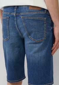 Shorts en denim de couleur bleue moyenne avec une coupe décontractée. Comprend cinq poches, des coutures visibles et un patch en cuir marron à l'arrière.