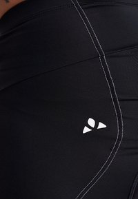 Leggings de sport noirs avec des coutures blanches contrastantes et un petit logo sur la cuisse. Le tissu semble lisse et extensible.
