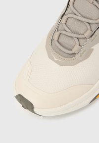 Αθλητικό παπούτσι σε ανοιχτό μπεζ χρώμα με επάνω μέρος με υφή κυψελώματος, γκρι λε πτομέρειες, ορατά κορδόνια και καουτσούκ σόλα με ελαφριά καμπύλη.