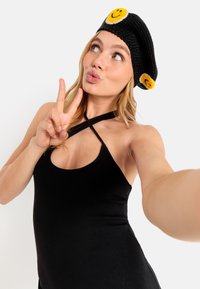 Schwarze gestrickte Baskenmütze mit zwei gelben Smiley-Gesichts-Applikationen. Das Modell trägt ein schwarzes ärmelloses Oberteil mit gerippter Textur.