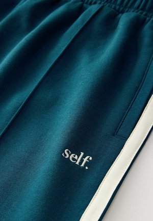 Türkiz színű melegítőnadrág, elasztikus derékrésszel. Fehér oldalsávokkal és a jobb lábon fehér hímzéssel ellátott "self." felirattal. Puha tapintású.