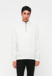 Un jeune homme aux cheveux courts et foncés, portant un sweat-shirt gris clair à fermeture zippée au quart et un pantalon noir, se tient devant un fond blanc.