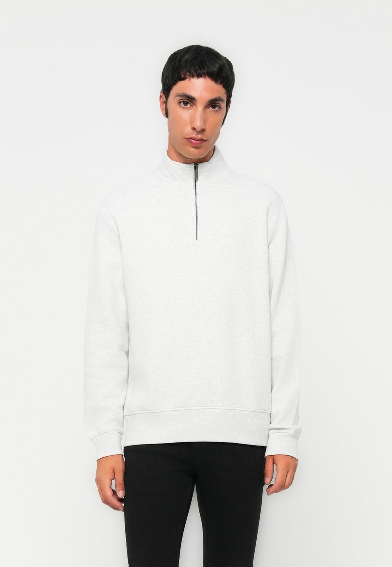 Un jeune homme aux cheveux courts et foncés, portant un sweat-shirt gris clair à fermeture zippée au quart et un pantalon noir, se tient devant un fond blanc.