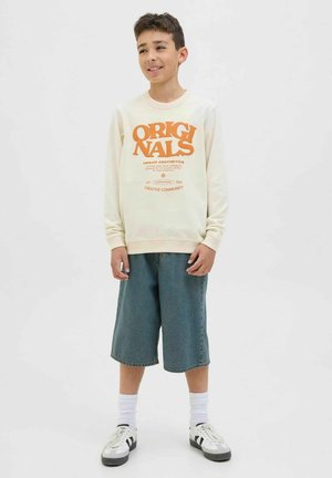 Cremefarbener Sweatshirt mit orangefarbener "ORIGINALS"-Aufschrift, kombiniert mit lockeren Jeansshorts. Sneakers mit schwarzen Akzenten, weiße Socken. Schlicher, lässiger Stil.