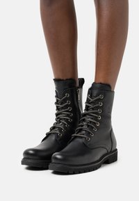 Bottes noires en cuir montant jusqu'à la cheville avec bord en fourrure, dotées d'œillets métalliques et de lacets épais et texturés. Semelles en caoutchouc robustes avec motif pour une meilleure adhérence.