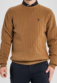 Man med en brun kabelstickad tröja över en svart skjorta med krage och mörka byxor, stående med händerna avslappnat vid sidorna.