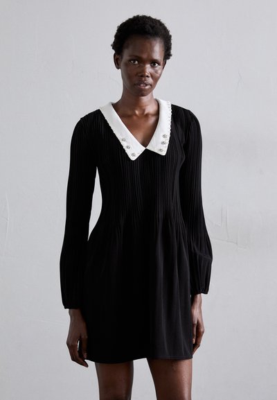 Vestido negro con plisados y mangas largas, que presenta un collar blanco adornado con pequeños botones decorativos. Textura suave y diseño ajustado.