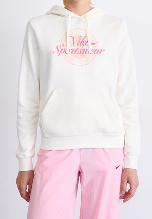 Osoba ubrana w białą bluzę Nike Sportswear z kapturem i kieszenią z przodu oraz jasnoróżowe spodnie z małym czerwonym logo Nike.
