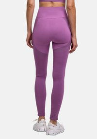 Frau in lila hochgeschnittenen Leggings und weißen Sportschuhen, die mit dem Rücken zu einem schlichten Hintergrund steht.