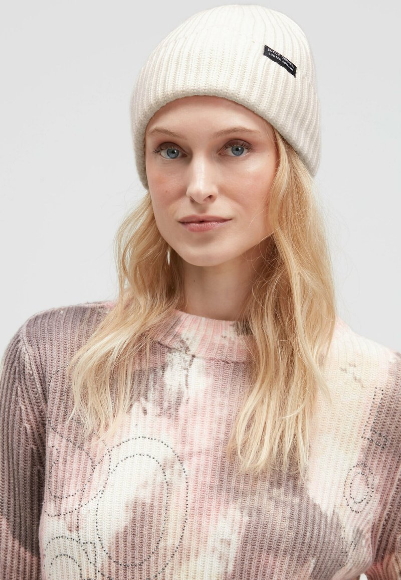 Beige ribbed knit beanie with a sewn label, worn with a multicolored, patterned sweater featuring a round neckline and textured fabric.