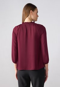 Vinrød bluse med trekvart ermer, med en volang halslinning og glatt, lett stoff. Kombinert med svarte bukser.