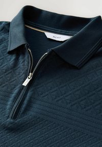 Polo de culoare albastru închis, cu închidere prin fermoar, având un model geometric texturat și un guler clasic. Eticheta vizibilă: NEXT.
