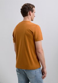 Bărbat cu păr scurt și creț, purtând un tricou portocalie cu mânecă scurtă și blugi de culoare albastru deschis, întors cu fața către un fundal gri simplu.