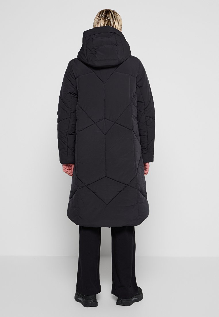 Langer schwarzer Puffermantel mit Kapuze, mit einem geometrischen gesteppten Design und einem mittellangen Schnitt, kombiniert mit schwarzen Hosen und Schuhen.