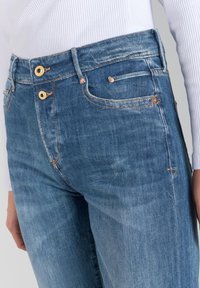 Un jean en denim de lavage bleu moyen avec une coupe droite. Comprend des boutons en cuivre, une petite poche à monnaie et un léger délavage sur le tissu.