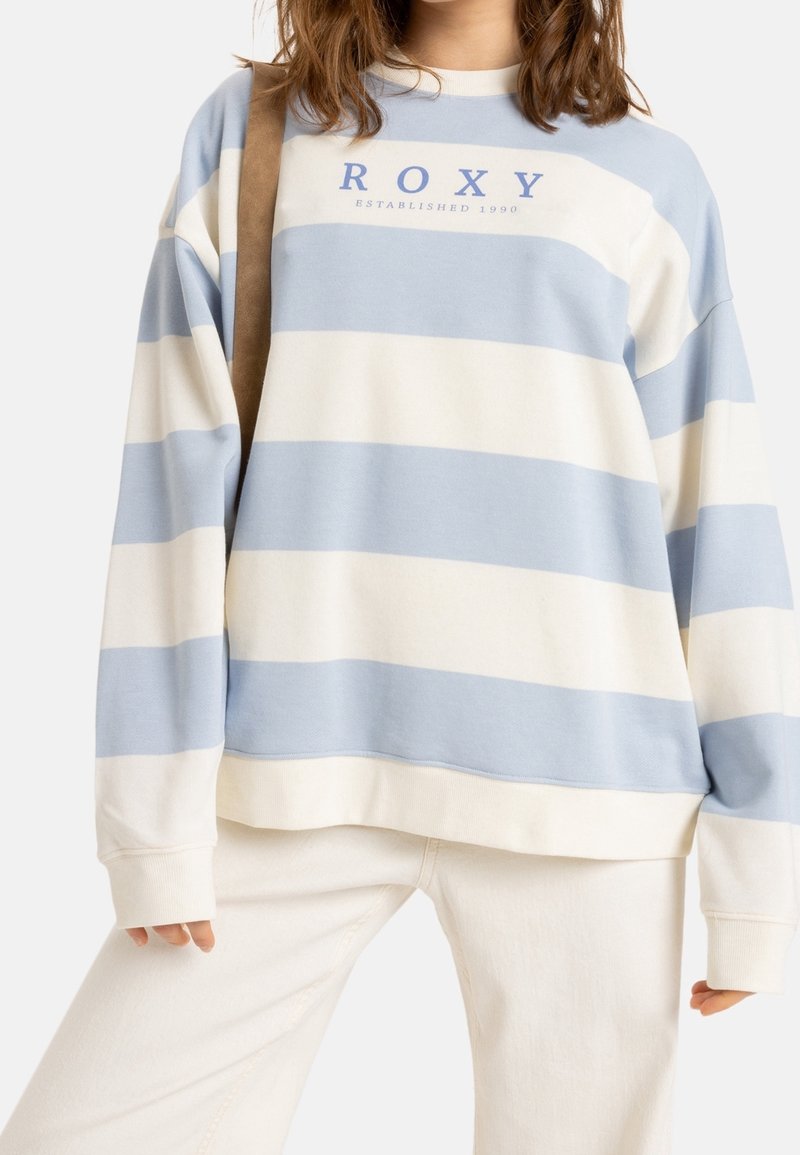Personne portant un sweat-shirt Roxy rayé bleu et blanc avec un pantalon beige et un sac à bandoulière marron, cadrée au niveau du cou.