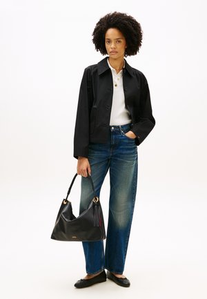 Donna con capelli ricci che indossa una giacca nera, una maglietta polo bianca, jeans blu, scarpe basse nere, che tiene una borsa nera con nappina.