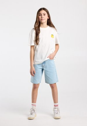 Meisje met lang bruin haar, gekleed in een wit T-shirt met een citroengrafiek, lichtblauwe denimshorts, witte sokken met rode strepen en witte sneakers.