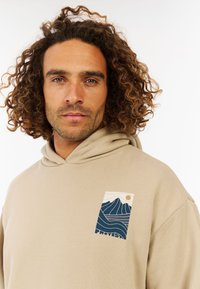 Beige hoodie met een voorzak, ontworpen met blauwe golfpatronen en een zon. Gemaakt van zachte stof met een ontspannen pasvorm.