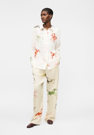 Γυναίκα που φοράει λευκό μπλουζάκι και παντελόνι με πολύχρωμα σχέδια αλόγων, στέκεται με τα χέρια στις τσέπες και καφέ παπούτσια.