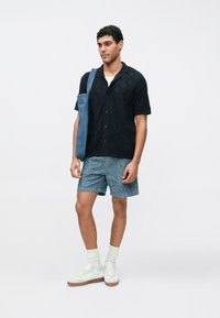 Zwart T-shirt met korte mouwen en een gestructureerd diamantpatroon, blauwe denim shorts, witte sneakers en lichtgekleurde sokken. Casual outfit met een tote bag.
