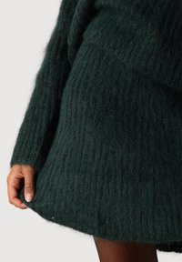 Nærbilde av en person som har på seg en mørkegrønn, fluffy strikket kjole, som holder kanten med den ene hånden mot en lys bakgrunn.