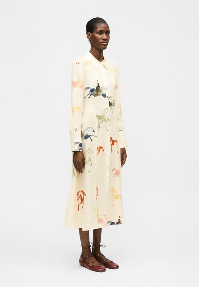 Γυναίκα με σκουρόχρωμο δέρμα φοράει μπεζ midi φόρεμα με μακριά μανίκια και πολύχρωμα σχέδια με άλογα, καθώς και μπορντό παπούτσια με κορδόνια.