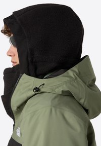 Osoba ubrana w zieloną i czarną kurtkę North Face z czarnym kapturem z polaru i szarymi okularami przeciwsłonecznymi, przedstawiona w lewym profilu.