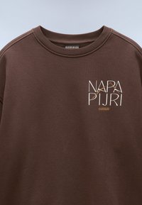 Brun bomullströja med rund hals, prydd med en grafisk logotyp i vitt och guld: "NAPA PIJRI" med geometriska accenter. Slät textur.