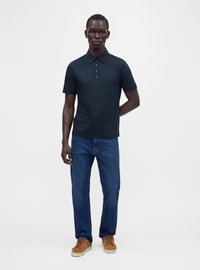 Homme portant un polo noir à manches courtes, un jean bleu et des baskets marron, debout devant un fond blanc uni.
