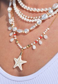Des colliers de perles superposés en blanc et or, présentant diverses formes et tailles de perles, agrémentés d'un pendentif étoile de mer.