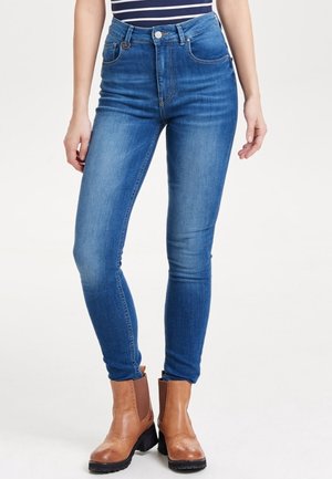 Kvinde iført højtaljede blå skinny jeans og lysebrune læderstøvler, stående mod en ensfarvet hvid baggrund.