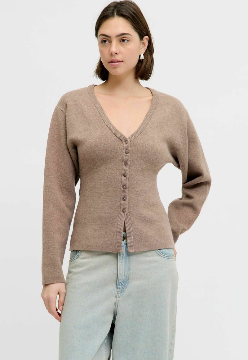 Vrouw draagt een taupekleurige knoopjesvest met V-hals en lange mouwen, gecombineerd met lichtblauwe jeans met hoge taille, staand met één hand op haar heup.