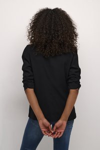 Osoba s kudrnatými vlasy, oblečená v černé bundě a modrých džínách, stojí zády k divákovi s rukama sepjatýma za zády, na jednobarevném pozadí.