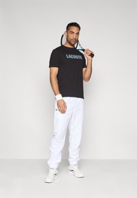 Czarna bawełniana koszulka z logo "LACOSTE", białe spodnie dresowe, białe buty sportowe i opaska na nadgarstek; zawodnik trzymający rakietę tenisową.