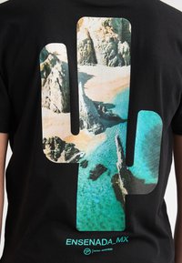 Camiseta de algodón negra con una gran silueta de cactus rellena con un paisaje de playa, con el texto "ENSENADA_MX" y un logo en turquesa.