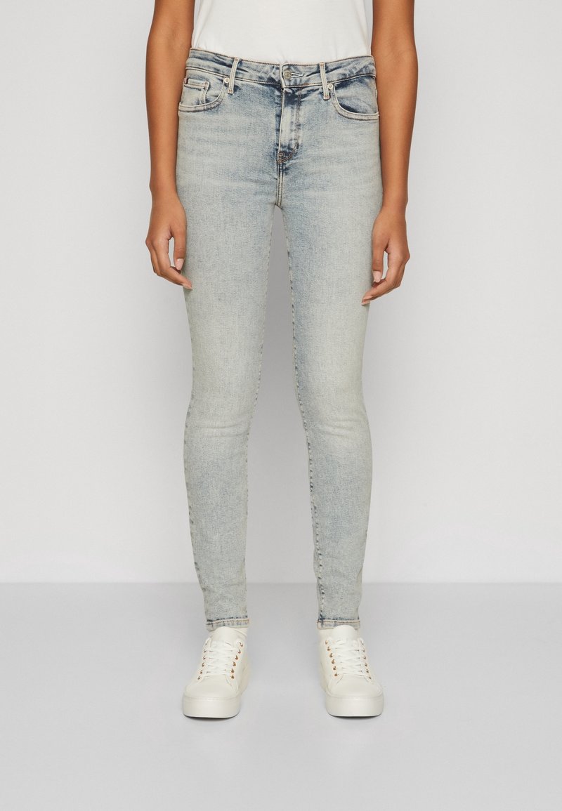 Jeans ajustados de color azul claro, elaborados en denim, con un clásico diseño de cinco bolsillos y un leve desteñido, combinados con zapatillas blancas.