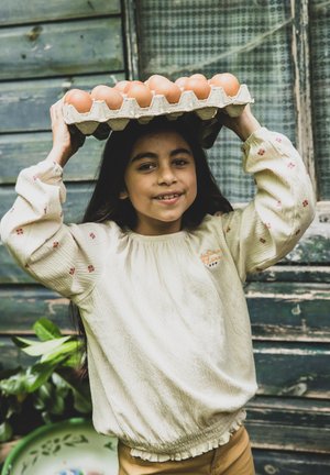 Een doos eieren op het hoofd van een meisje in een lichtbeige gebreide trui met bloemenaccenten. De achtergrond bestaat uit rustieke houten planken.