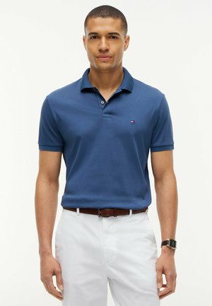 Mann trägt ein blaues Polohemd mit kleinem Logo, weiße Hose, braunen Gürtel und eine schwarze Armbanduhr vor hellem Hintergrund.