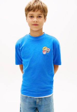 Ragazzo con capelli biondi corti che indossa una maglietta blu con piccole toppe colorate a forma di frutta sul petto e jeans azzurri.