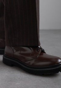 Chaussures en cuir marron à texture lisse, à bouts ronds et avec des lacets. Semelles en caoutchouc à motif strié, associées à un pantalon marron à rayures.