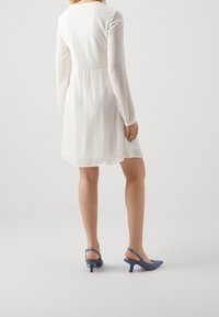 Vit långärmad klänning med en utsvängd nederkant, tillsammans med blå slingback-hälar med spetsiga tår och en liten häl. Slät textur.